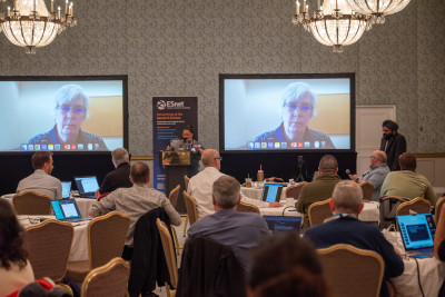 View from back of conference room toward presentation screen. Room is full of circular tables with people sitting around them. On screen is the face of a person speakering.