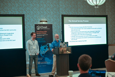 Two people are standing at the front of a large room by a podium. An ESnet banner and large presentation screens are behind them. Presentation screen title is The Annual Survey Process
