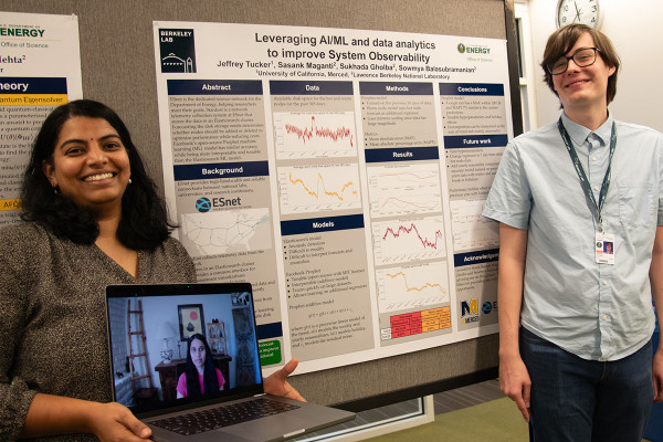 Woman holding a laptop with woman on zoom, standing next to student and a poster