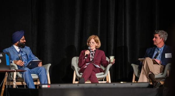 Carol Burns speaks into a microphone while Monga and Serrao look in